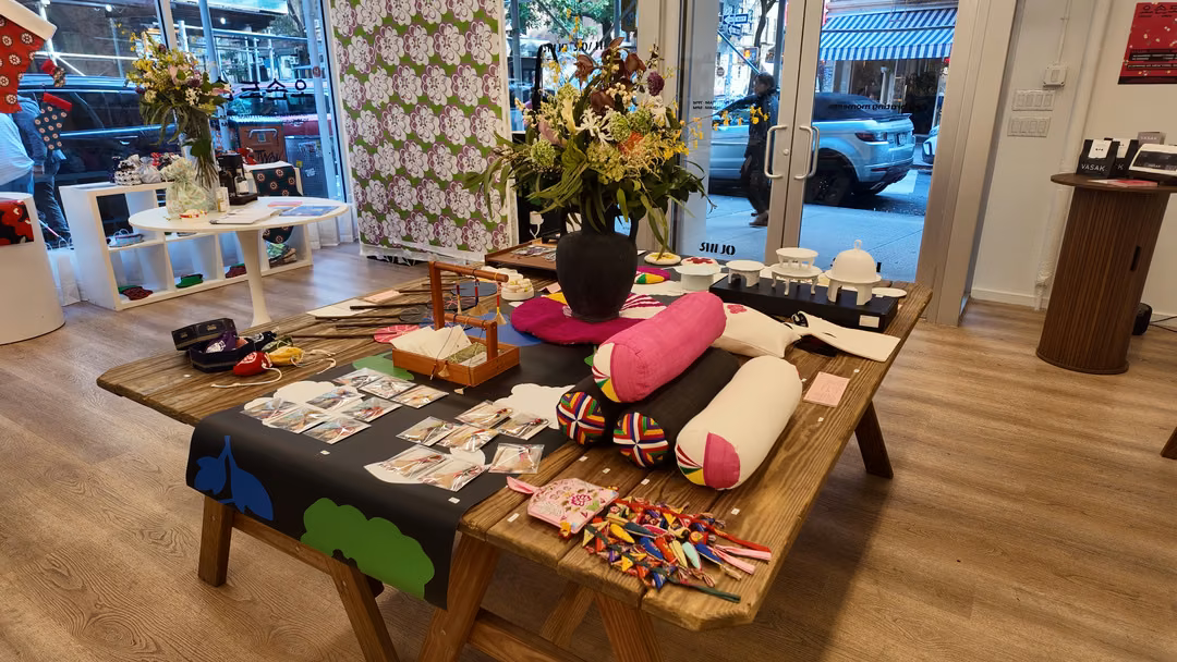 A wooden display table with a black cloth, pink and cream bolster cushions, white tableware, a vase of flowers and small accessories, in front of the shop’s street door