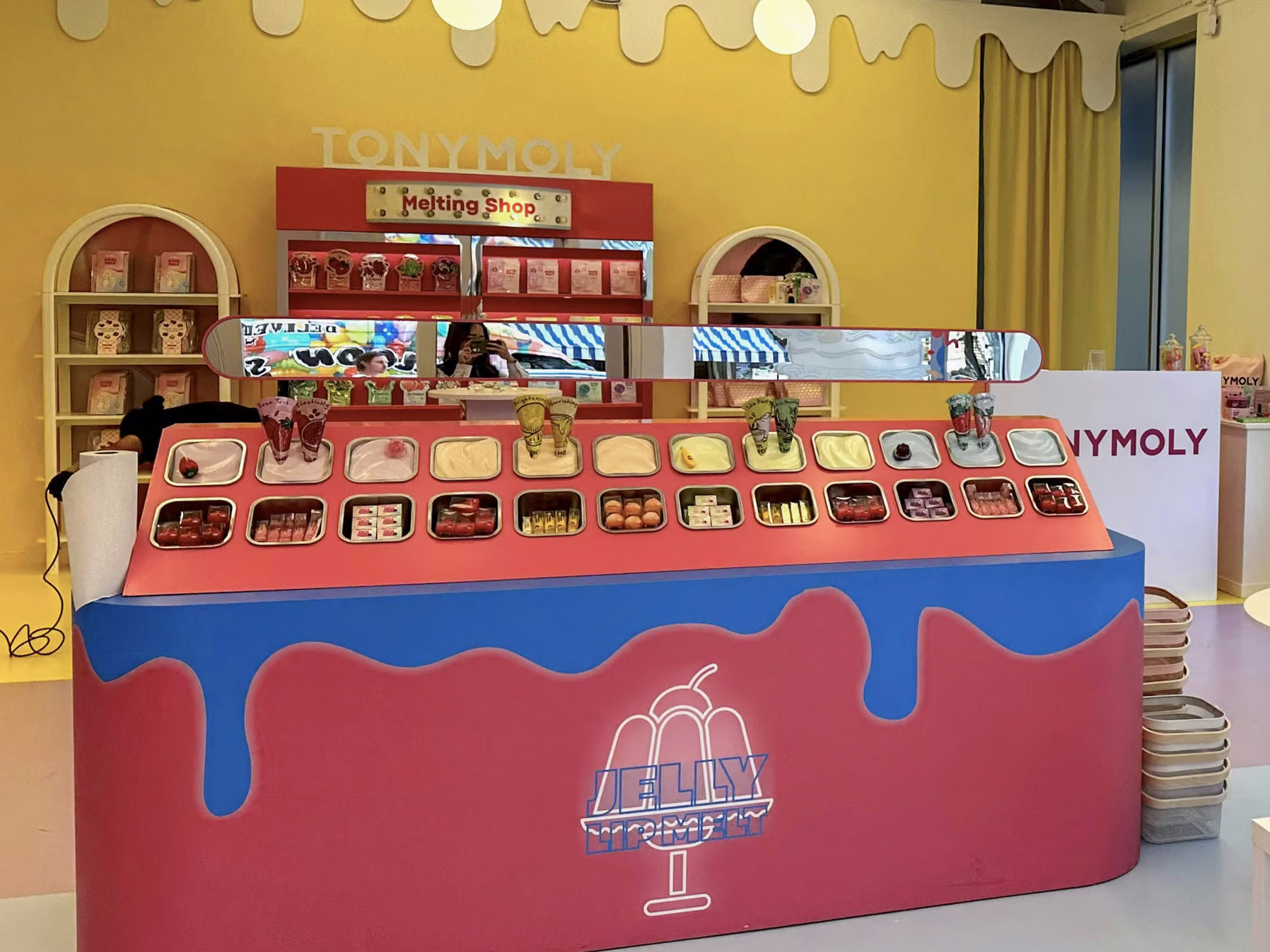 The TONYMOLY Melting Shop pop-up in New York: a red counter with a blue drip, trays of lip products and a bright yellow wall